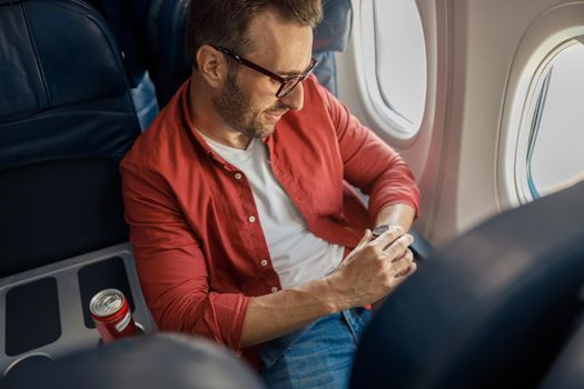 Handsome caucasian man in casual wear and glasses checking arrival time on his watch, sitting on the plane near the window. Relax, travel, vacation, transportation concept