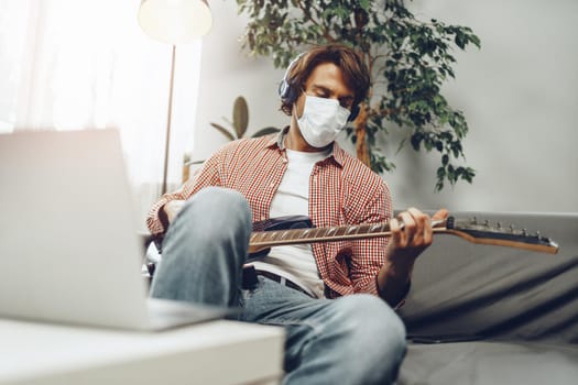 Young man plays guitar at home in medical mask. Coronavirus quarantine and self-isolation lifestyle concept