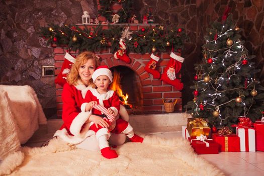 Mother and son are sitting near fireplace and christmas tree with gift boxes.