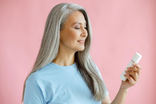 Happy middle aged hoary haired Asian woman looks bottle of cosmetic product on pink background in studio. Mature beauty lifestyle