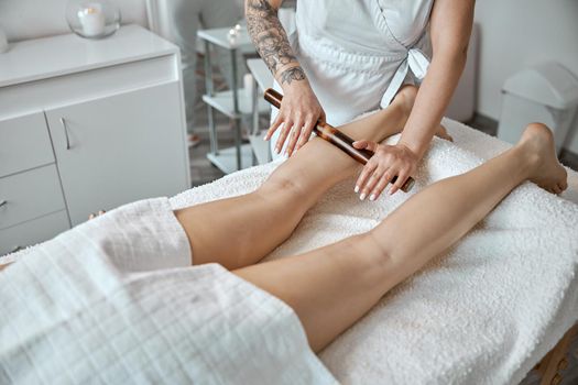 Conident female specialist is doing leg massage with bamboo stick to beautiful caucasean woman client