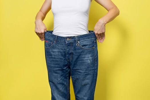 The concept of diet, proper nutrition, weight loss. Slim Woman Showing Loose Jeans and her Loss Weight. Woman white t-shirt and oversize jeans isolated on yellow background.