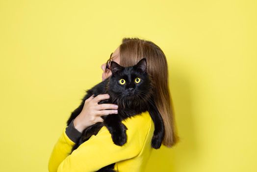 Girl in a yellow dress holds her beloved fluffy black cat with yellow eyes on a yellow background. Pets care concept.