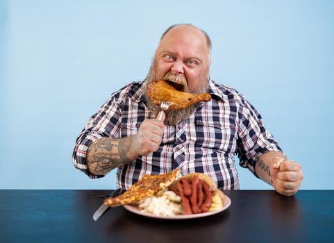 Funny bearded man with overweight eats smoked chicken leg at table with rich food on light blue background in studio