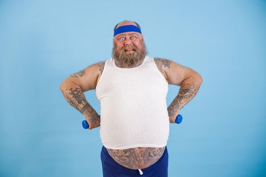 Happy bearded plump man with small dumbbells holds hands on waist posing on light blue background in studio