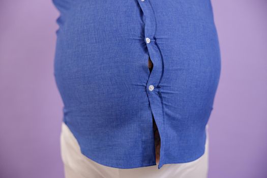 Man with overweight and tattoos on arms in tight blue shirt holds hands on large belly standing on purple background in studio closeup