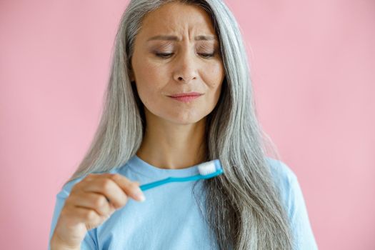 Upset hoary haired mature Asian lady in blue t-shirt looks at toothbrush posing on pink background in studio. Oral cavity hygiene