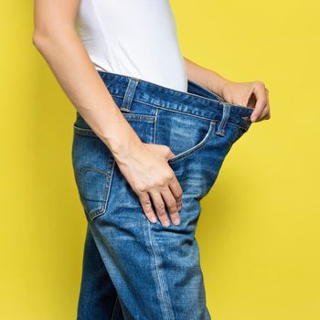 The concept of diet, proper nutrition, weight loss. Slim Woman Showing Loose Jeans and her Loss Weight. Woman white t-shirt and oversize jeans isolated on yellow background.
