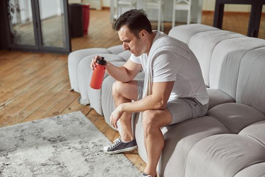 Top view of thirsty man with botle of water and towel sitting on sofa after workout at home