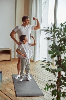 Happy muscular man and boy are standing on mat and showing biceps during domestic workout