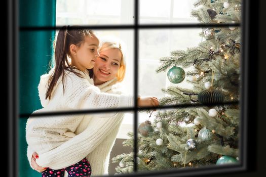 Young big family celebrating Christmas enjoying dinner, view from outside through a window into a decorated living room with tree and candle lights, happy parents eating with three kids