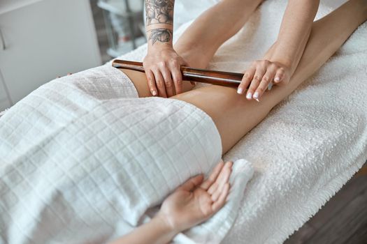 Conident female specialist is doing leg massage with bamboo stick to beautiful caucasean woman client