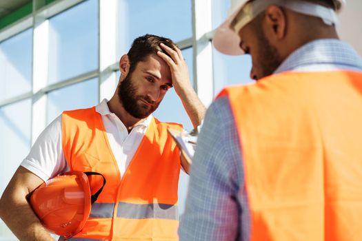 Two young male engineers in uniform and hardhats working at construction site, close up