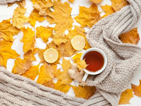 mug of tea with yellow maple autumn leaves and lemons with ginger. autumn seasonal flat lay hot drink composition. grey scarf as autumn background.