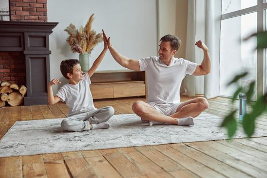 Happy sporty boy is training with dad together on mat at home and they are motivating each other