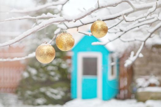 golden christmas tree balls on tree branches and winter snow. Christmas balls near fur-trees covered with a snow and small wooden house. Playground for children at backyard