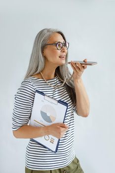 Pretty mature Asian businesswoman in casual clothes records audio message on cellphone holding diagrams on light grey background in studio