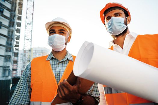 Two men in workwear and medical masks working with blueprints on object, close up