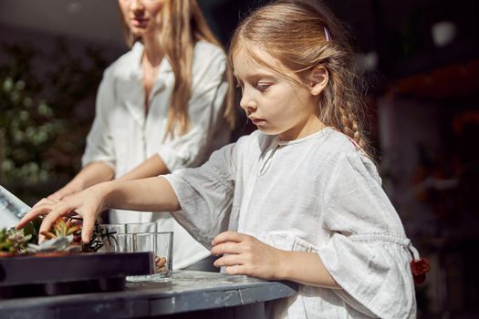 caucasian confident happy florist is working with her young daughter and making composition from glass stones and plants in botanic shop