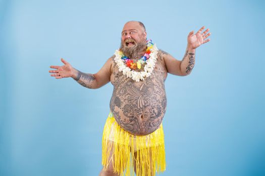 Positive bearded fat man with different tattoos in decorative yellow grass skirt and flowers garlands dances on light blue background in studio