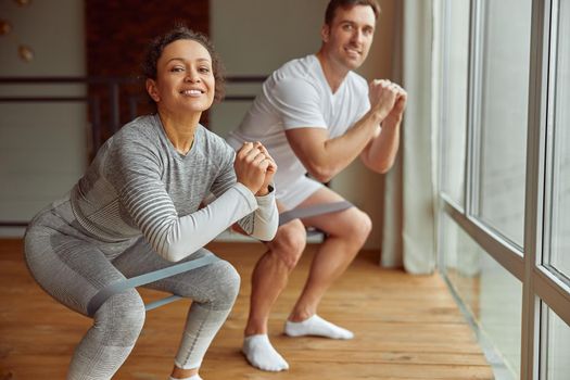 Full length portrait of jolly sporty man and woman doing training with resistance bands before window