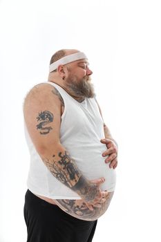Happy fat man with big belly and tattoes in sports wear is holding his stomach
