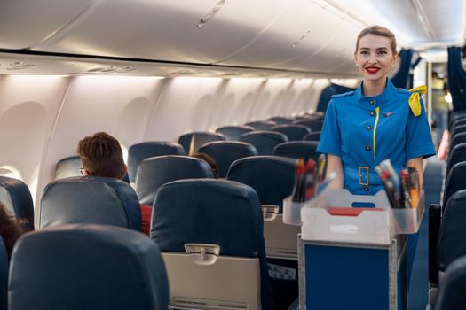 Smiling stewardess serving food to passengers on aircraft. Air hostess walking with trolley on aisle. Travel, service, transportation, airplane concept