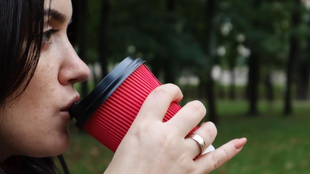 Young stylish woman drinks morning hot coffee in red eco paper cup outdoors in autumn park. Close-up side view of young woman holding takeaway coffee cup