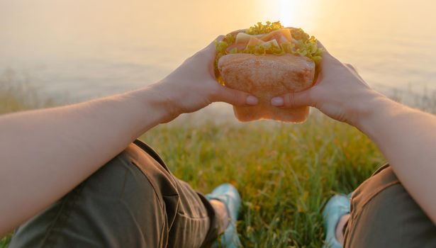 Woman holding fresh tasty burger sandwich and resting on summer coastline in front of sea outdoor, point of view.