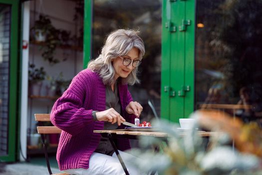 Happy mature woman with glasses in purple jacket eats delicious dessert at table on outdoors cafe terrace on autumn day