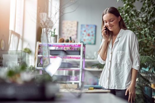 Confident female florist is working with dryed flowers in cozy flower shop while speaking on the smartphone