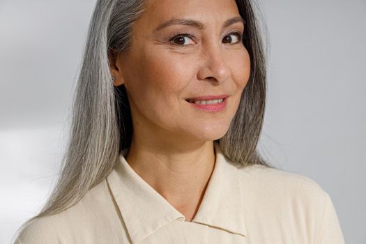 Attractive grey haired lady in stylish shirt smiles to camera on light background in studio. Mature beauty lifestyle