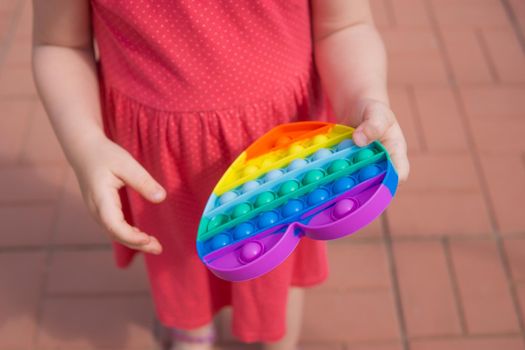 Children's hands hold a silicone toy. Push Popper fidget toy is the ideal toy to get rid of your frustration or stress. Kid having fun with popular simple dimple multicolored rainbow colors toy. Kids being kids LGBTQ colors Vertical. Summer vibes. Outdoor Outside.
