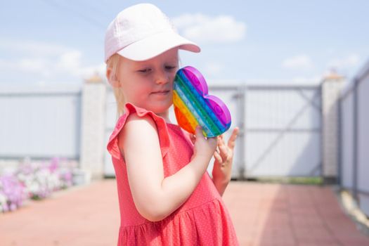 Cute caucasian fair hair girl in pink dress is playing and showing funny trendy silicone antistress colorful toy popit. Push Popper fidget toy is the ideal toy to get rid of your frustration or stress.