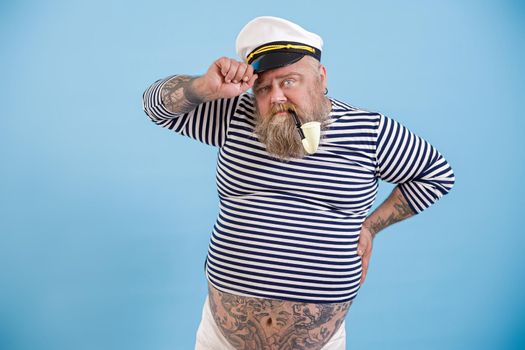 Mature fat sailor with beard and vintage smoking pipe adjusts hat looks into camera on light blue background in studio