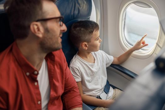 Curious little boy looking out the window while traveling by plane together with his father. Family, transportation, vacation concept