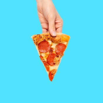 Slice of pepperoni pizza in hand isolated on blue. Top view on paperoni pizza. Concept for italian food, street food, fast food, quick bite