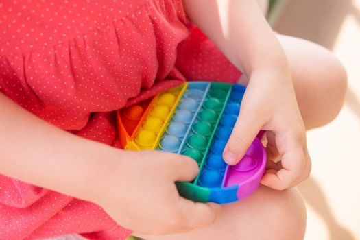 Cute caucasian fair hair girl in pink dress is playing and showing funny trendy silicone antistress colorful toy popit. Push Popper fidget toy is the ideal toy to get rid of your frustration or stress. Kid having fun with popular simple dimple multicolored rainbow colors toy. Children's hands hold a silicone toy. Kids being kids LGBTQ colors Vertical. Summer vibes. Outdoor Outside.