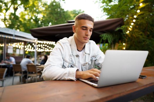 Young man sitting at table and typing on laptop keyboard while working in outdoor cafe, close up