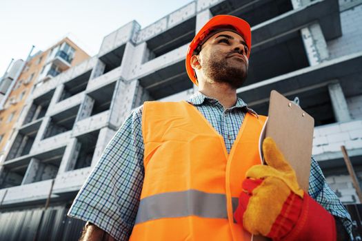 Young man engineer in workwear standing in construction site with clipboard, close up portrait