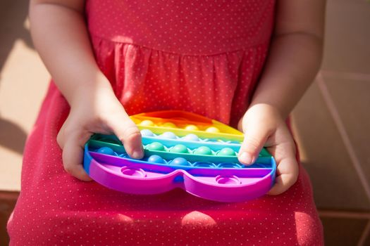 Cute caucasian fair hair girl in pink dress is playing and showing funny trendy silicone antistress colorful toy popit. Push Popper fidget toy is the ideal toy to get rid of your frustration or stress. Kid having fun with popular simple dimple multicolored rainbow colors toy. Children's hands hold a silicone toy. Kids being kids LGBTQ colors Vertical. Summer vibes. Outdoor Outside.