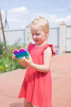 Cute caucasian fair hair girl in pink dress is playing and showing funny trendy silicone antistress colorful toy popit. Push Popper fidget toy is the ideal toy to get rid of your frustration or stress. Kids being kids LGBTQ colors Vertical