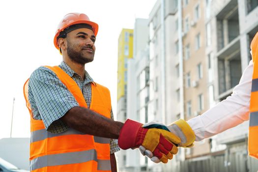 Two men engineers in workwear shaking hands against construction site, close up