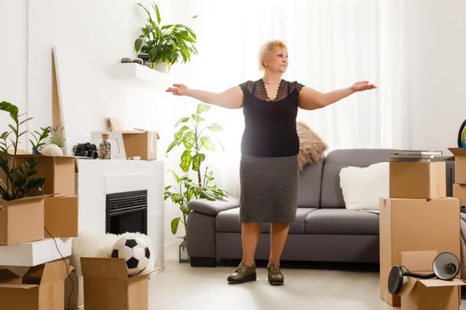 Happy excited mature retired middle aged woman standing in modern living room interior alone with arms outstretched enjoy freedom and wellbeing feel motivated at new furnished renovated home concept
