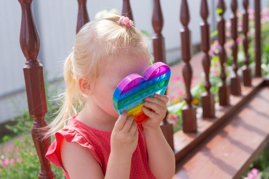 Cute caucasian fair hair girl in pink dress is playing and showing funny trendy silicone antistress colorful toy popit. Push Popper fidget toy is the ideal toy to get rid of your frustration or stress.