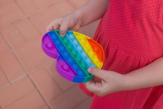 Children's hands hold a silicone toy. Push Popper fidget toy is the ideal toy to get rid of your frustration or stress. Kid having fun with popular simple dimple multicolored rainbow colors toy. Kids being kids LGBTQ colors Vertical. Summer vibes. Outdoor Outside.