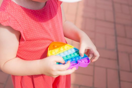 Cute caucasian fair hair girl in pink dress is playing and showing funny trendy silicone antistress colorful toy popit. Kid having fun with popular simple dimple multicolored rainbow colors toy. Children's hands hold a silicone toy. Kids being kids LGBTQ colors Vertical.