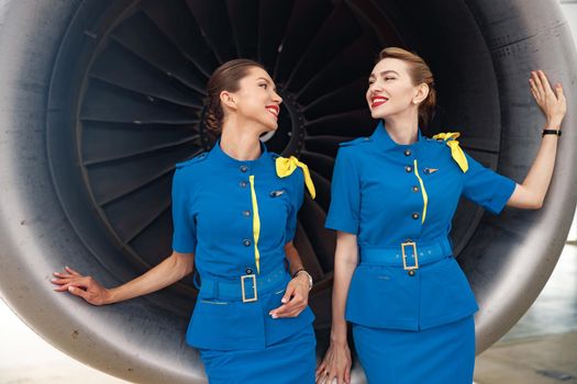 Two pretty air stewardesses in stylish blue uniform smiling at each other, standing together in front of aircraft engine. Occupation concept