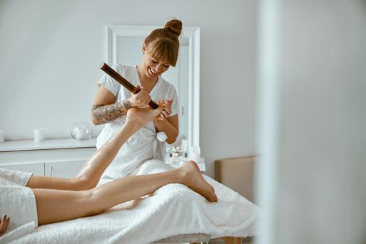 Conident female specialist is doing leg massage with bamboo stick to beautiful caucasean woman client