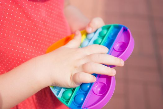 Cute caucasian fair hair girl in pink dress is playing and showing funny trendy silicone antistress colorful toy popit. Kid having fun with popular simple dimple multicolored rainbow colors toy. Children's hands hold a silicone toy. Kids being kids LGBTQ colors Vertical.